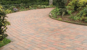 A brick paved driveway leads to a lush garden in the background, showcasing a blend of hardscape and greenery.